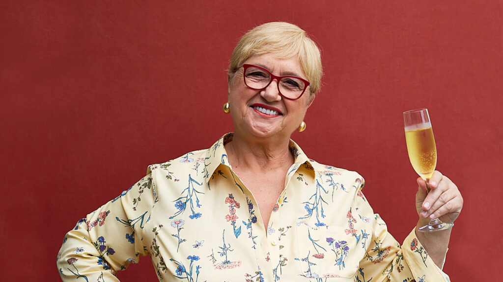 Lidia Bastianich in front of a red background holding a wine glass.