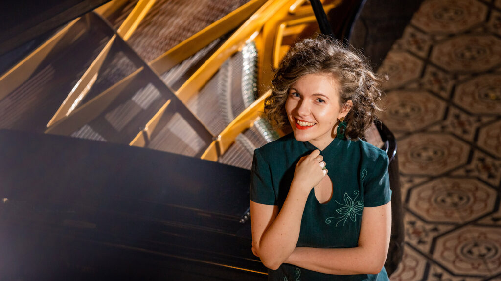 Woman smiling beside a grand piano with a patterned floor in the background.
