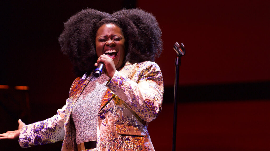 A person passionately singing into a microphone, wearing a colorful jacket and sparkling top, with large curly hair against a red background.