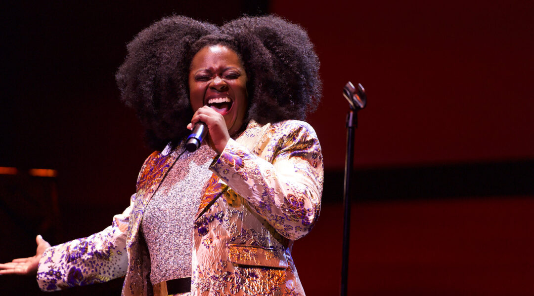 A person passionately singing into a microphone, wearing a colorful jacket and sparkling top, with large curly hair against a red background.