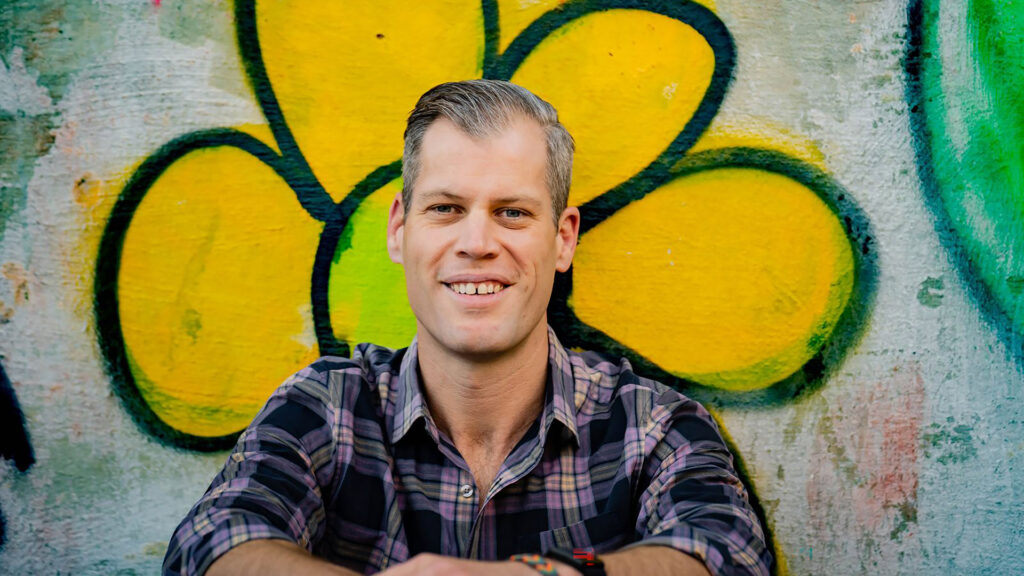 A person smiles in front of a graffiti mural depicting a large yellow flower.