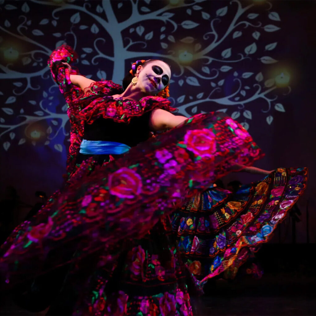 A dancer in a vibrant floral dress performs on stage with a decorative backdrop featuring a tree pattern.