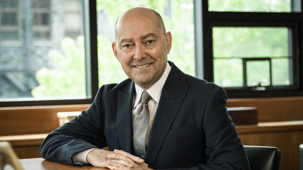 A bald man in a suit smiling while seated at a table in front of a window.