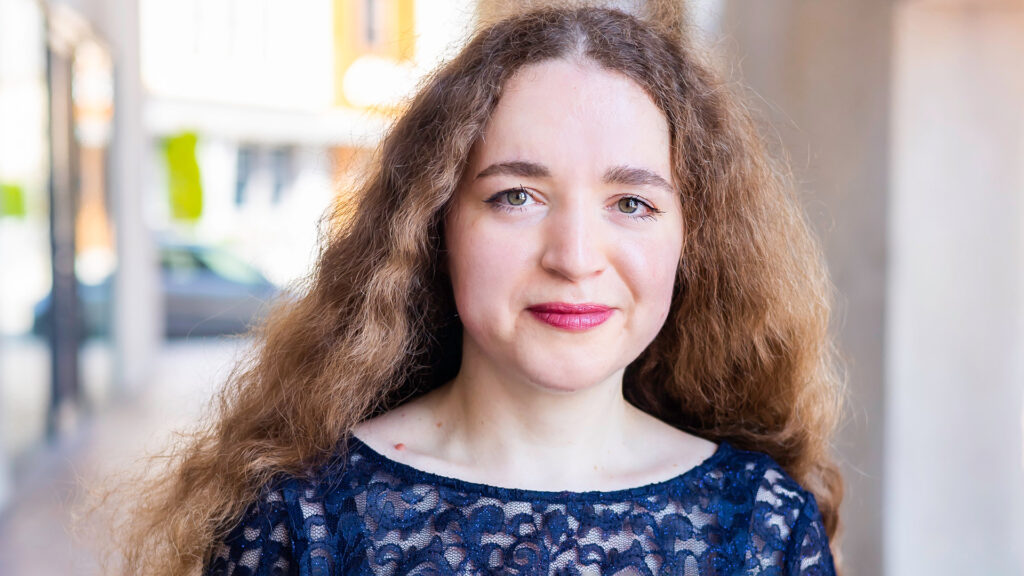 Person with long curly brown hair and a dark blue lace top outdoors with blurred urban background.