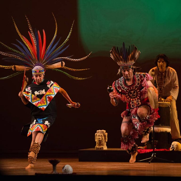 A traditional dance performance with two dancers in colorful outfits and feathered headdresses, accompanied by a drummer.