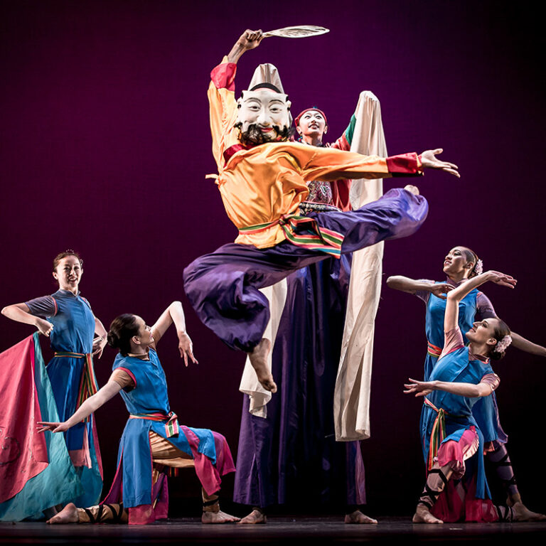 A dynamic dance performance with a masked dancer leaping in the center, surrounded by others in colorful costumes.
