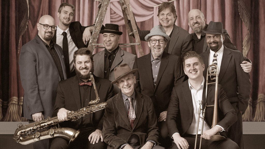 A sepia-toned group portrait of ten men in formal attire with musical instruments, in front of stage curtain backdrop.