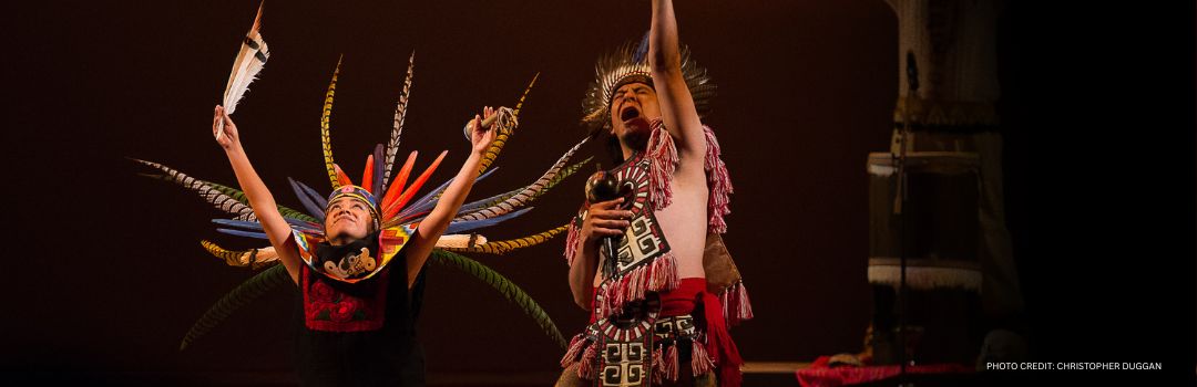 TREKKING MEXICO HEADER Two performers in vibrant traditional costumes with feathered headdresses on stage. PHOTO CREDIT: CHRISTOPHER DUGGAN
