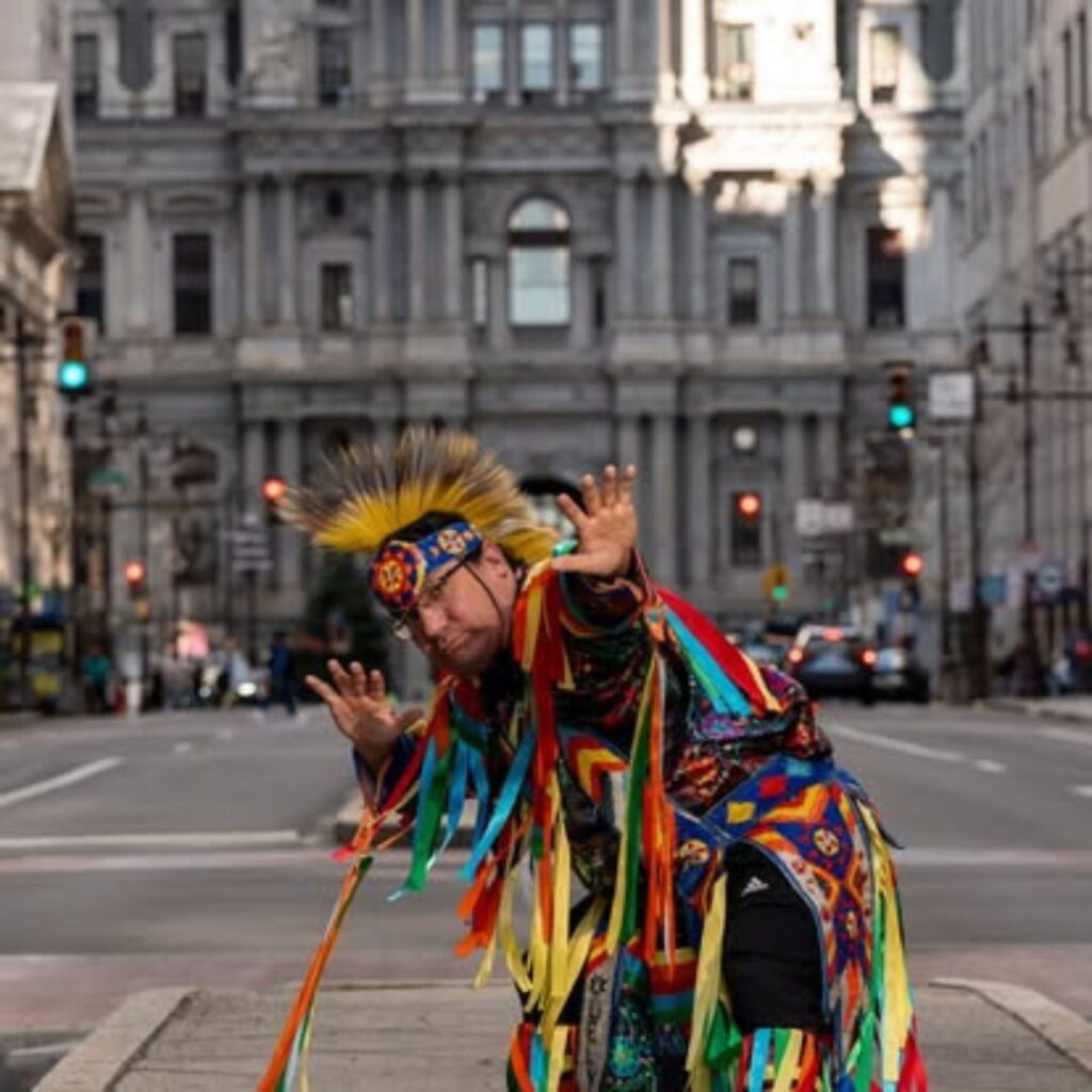 Person in colorful traditional attire poses on a city street with a historical building in the background.