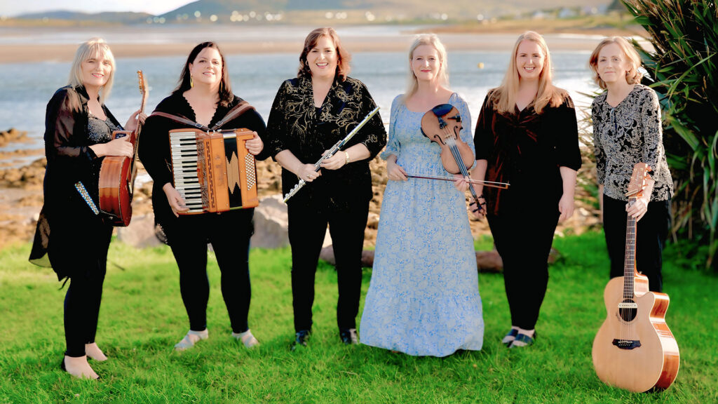 Six women with musical instruments standing on a grassy area by water.