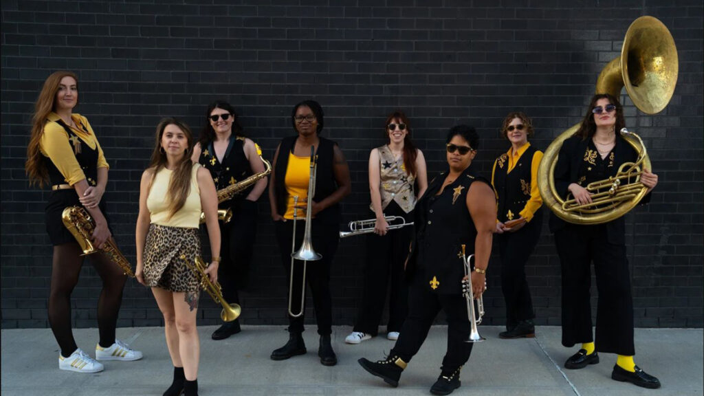 A group of eight people in coordinated black and gold outfits, holding various brass instruments, stand in front of a black brick wall.