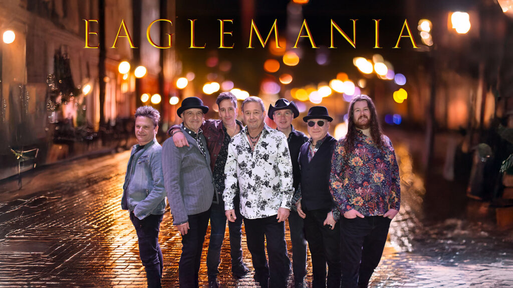 A group of seven men in varied attire standing on a dimly lit cobblestone street at night with blurred streetlights in the background.
