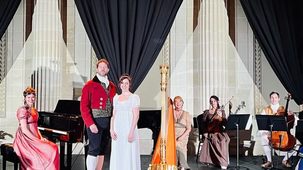 A group of six people in period costumes on a stage with musical instruments, dark curtains, and columns.