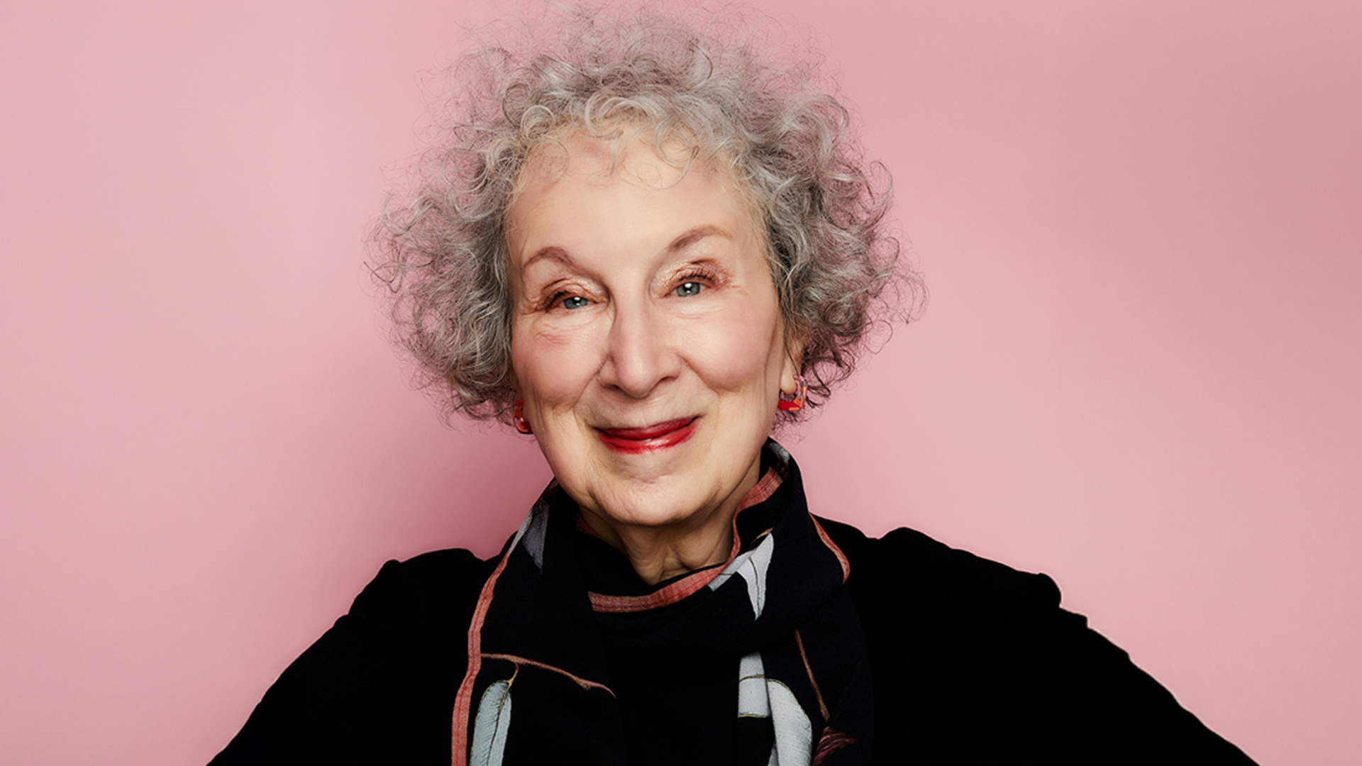 Margaret-Atwood—1920×1080 Person with curly gray hair smiling in front of a pink background.