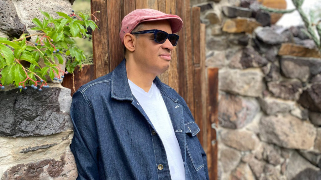 Person in a pink cap and denim jacket leaning against a stone wall with green leaves and blue berries nearby.