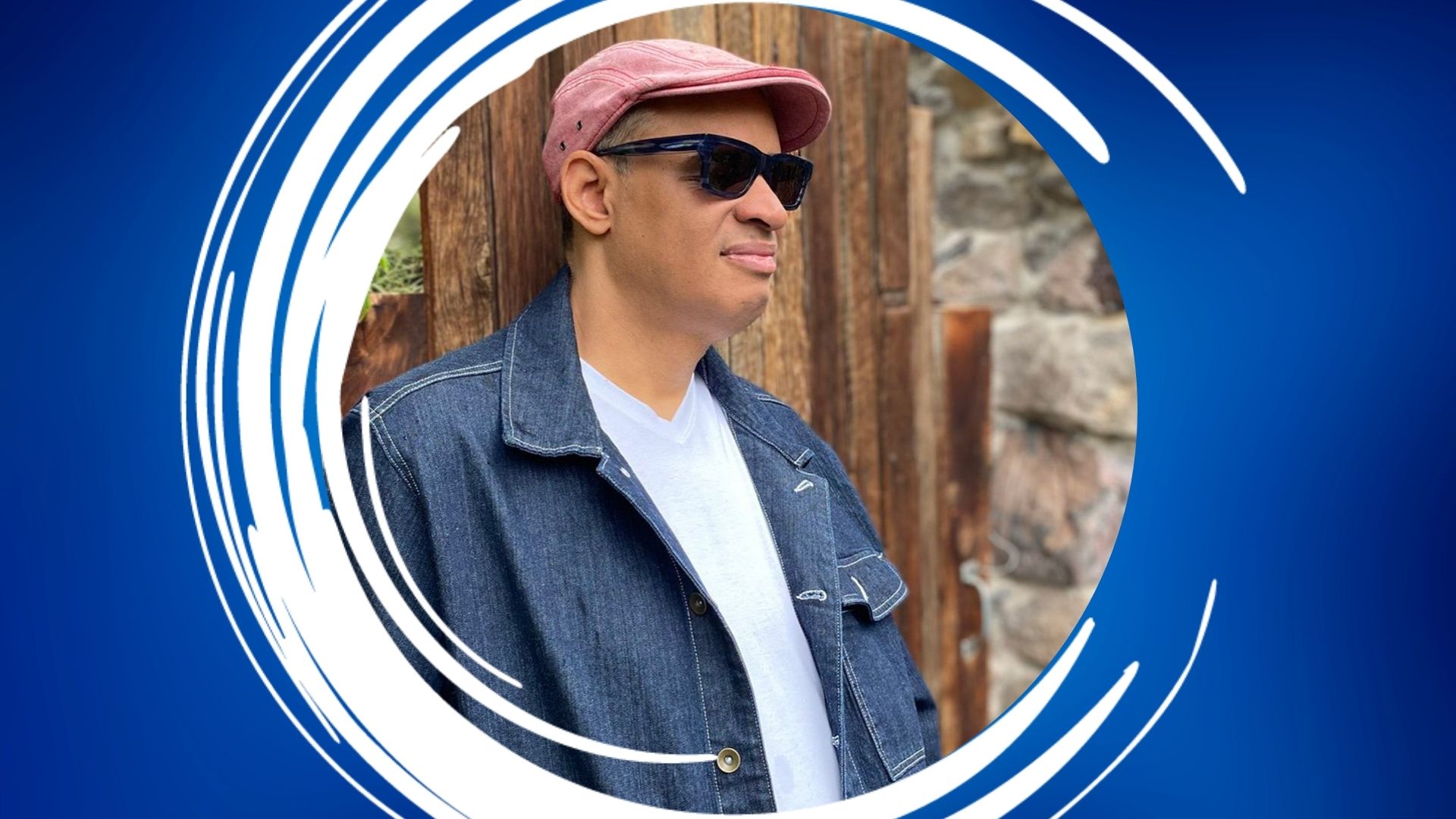 Raul Midon – 1920×1080 Feature Person wearing a pink flat cap and sunglasses in front of a wooden background.