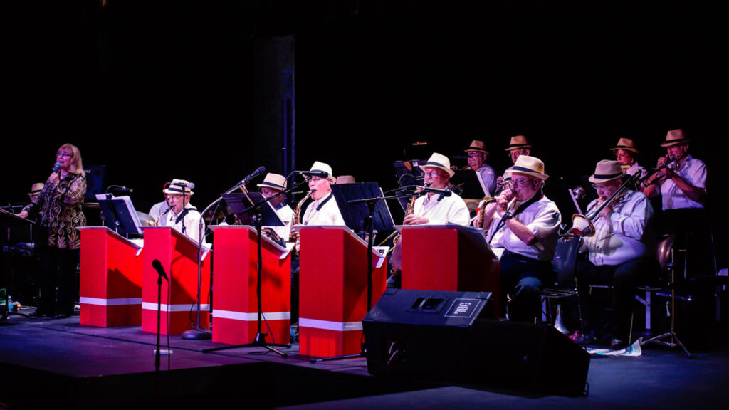 A jazz band performing on stage with bright red music stands and wearing white shirts and straw hats.