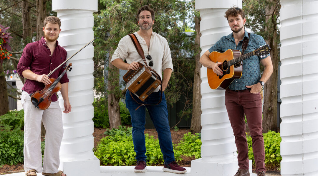 Three men with musical instruments standing between white columns surrounded by greenery.