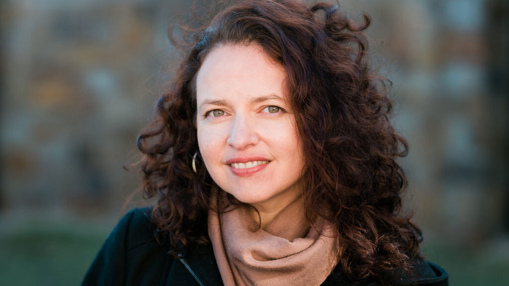 Portrait of a woman with curly dark hair, wearing a brown scarf and black coat, against a blurred background.