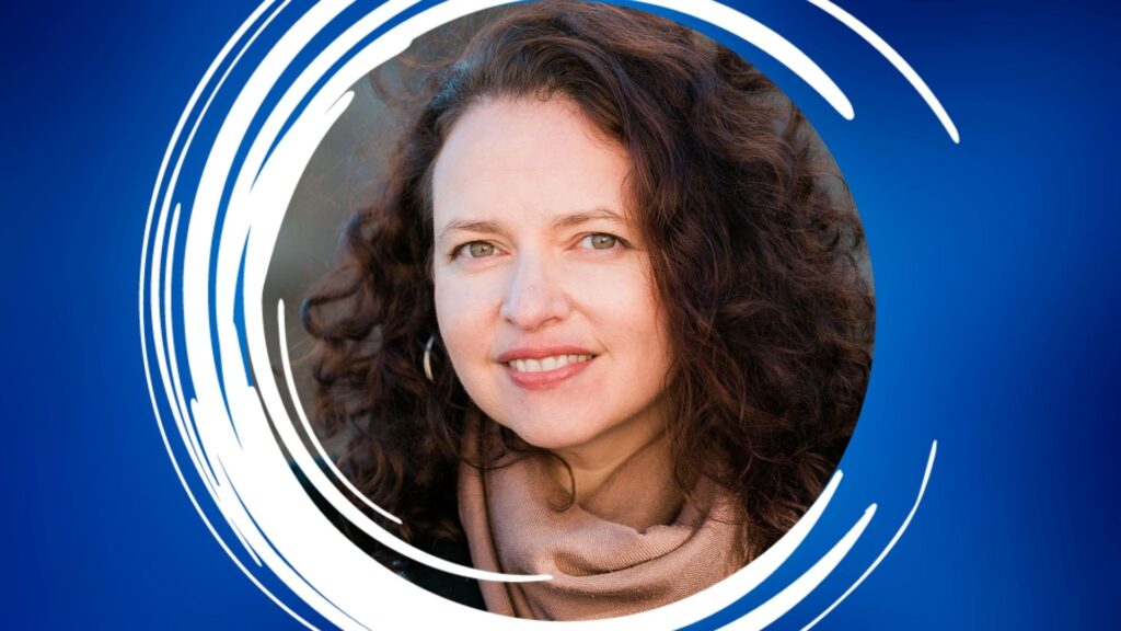 A person with curly dark brown hair, wearing a black coat and brown scarf, stands outside with a blurred stone wall in the background.
