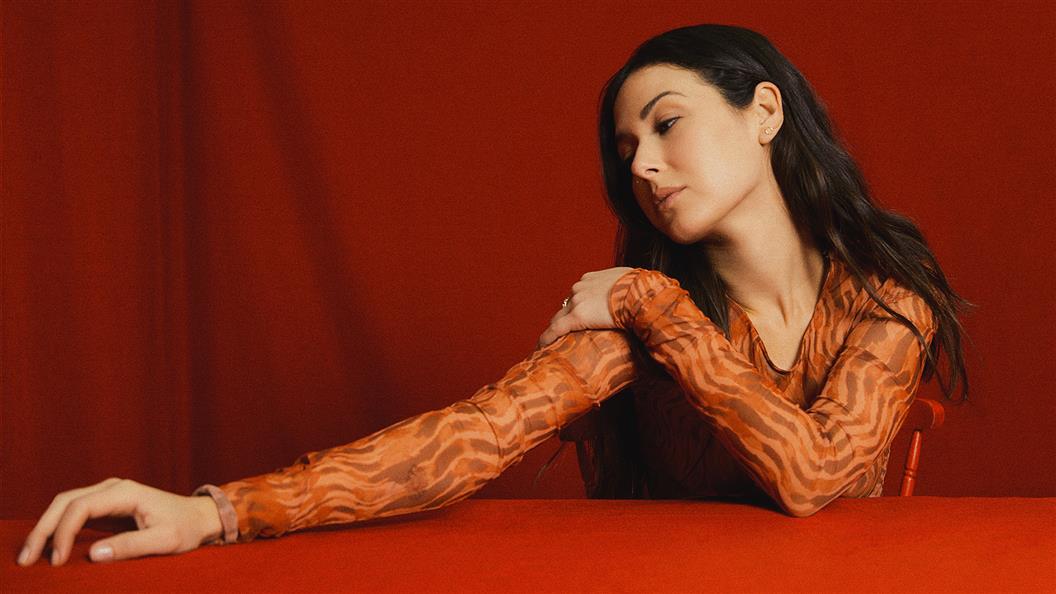 Woman in orange patterned top seated at a red table with red background.