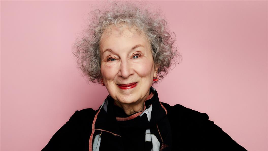 A white woman with grey curly hair wearing a black sweater and scarf in front of a pink backdrop.