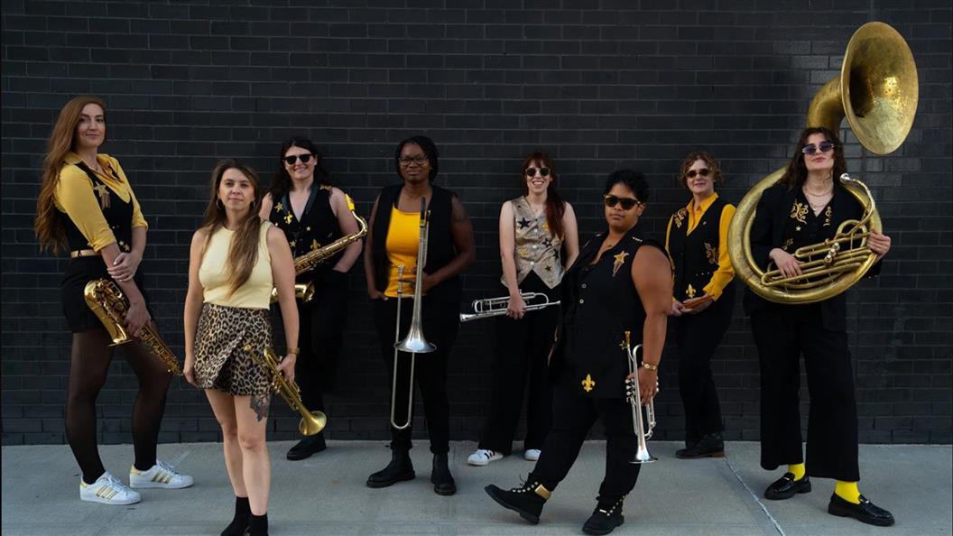 A group of eight musicians in black and yellow outfits holding brass instruments against a black brick wall.