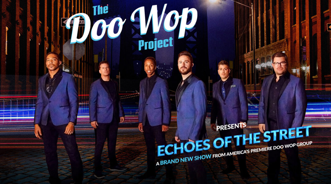 Six men in blue suits on a cobblestone street with streaks of light and surrounding buildings at night.