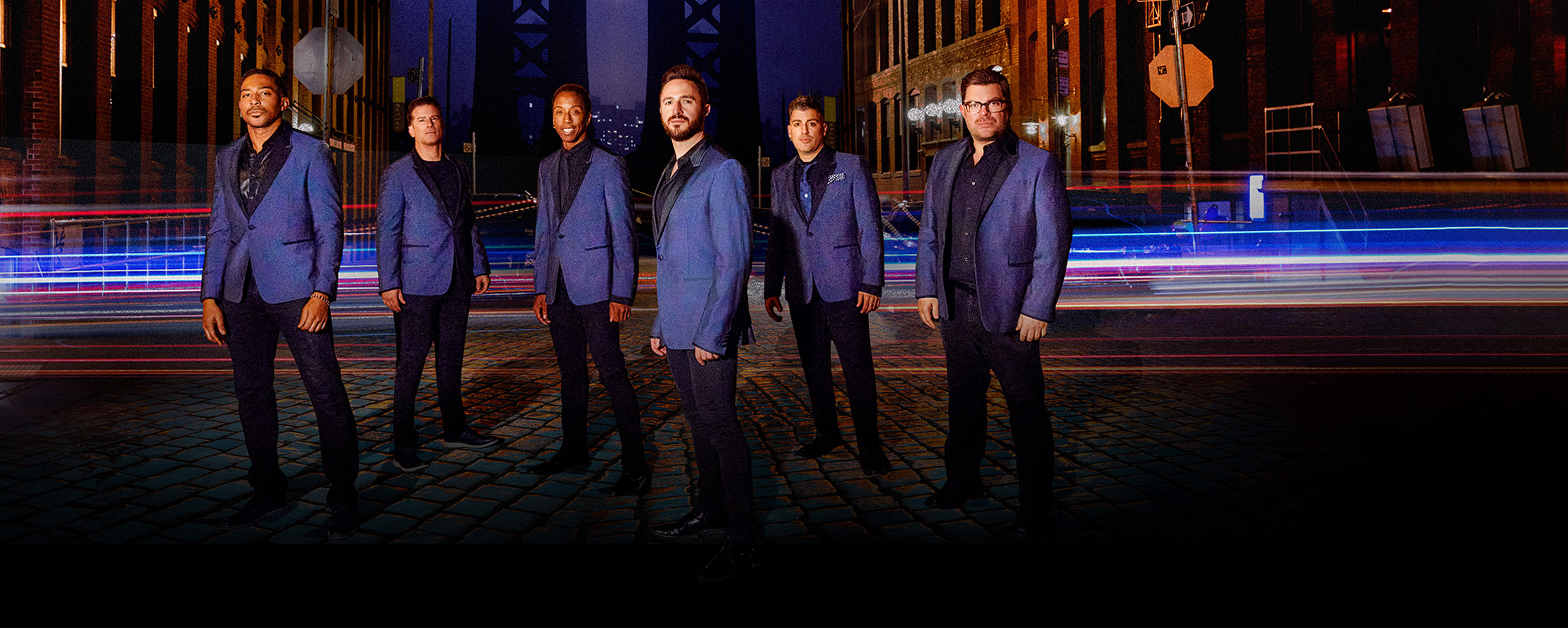 Six men in blue blazers stand on a cobblestone street at night, with city lights and vehicle trails behind them.
