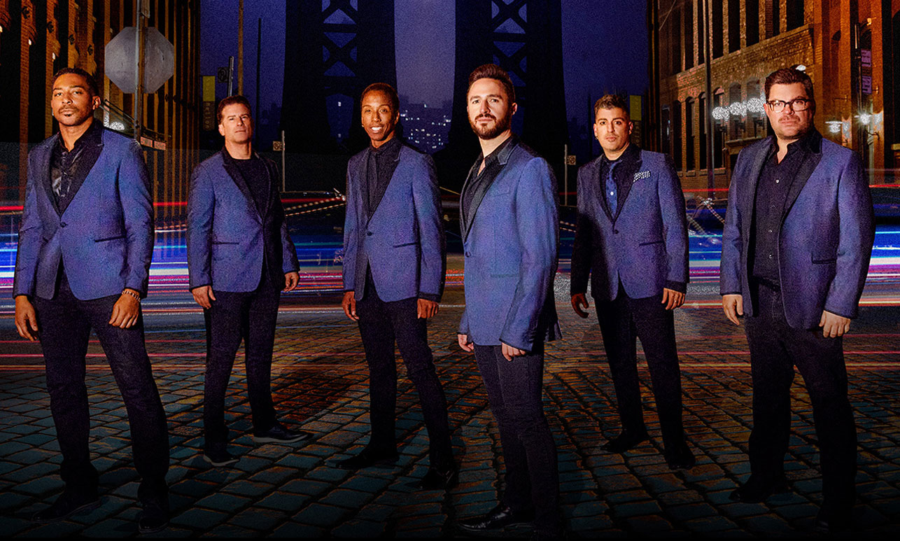 Six men in blue suits on a cobblestone street with streaks of light and surrounding buildings at night.