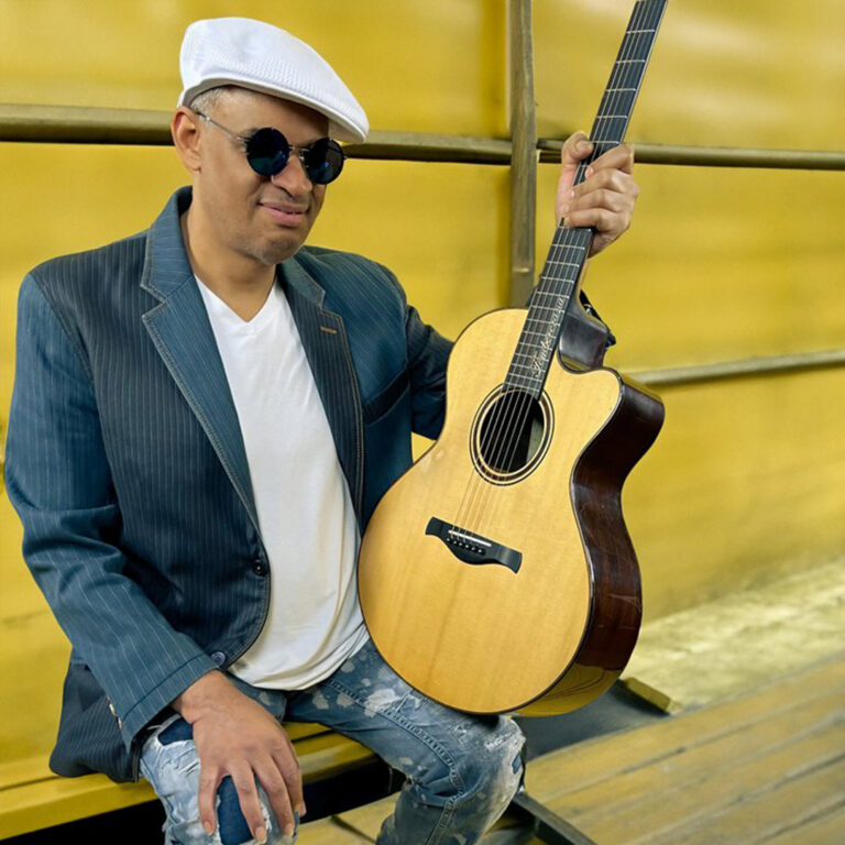 A man in a white had holding a guitar in front of a yellow wall