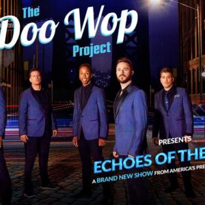 Six men in blue suits on a cobblestone street with streaks of light and surrounding buildings at night.