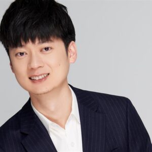 Person in a dark suit with a white shirt smiling against a gray background.