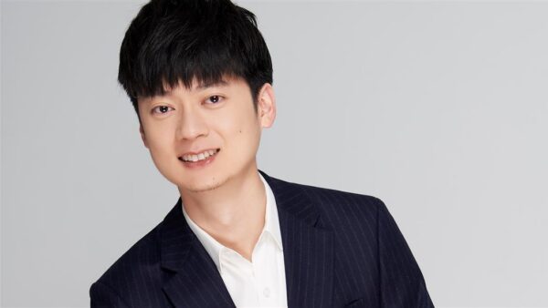 Person in a dark suit with a white shirt smiling against a gray background.