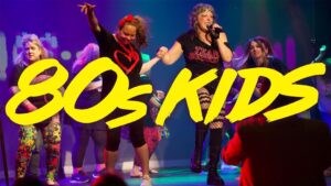 A lively performance with people in 80s-themed clothing, illuminated by colorful stage lights, with the text "80s KIDS" in bold yellow.