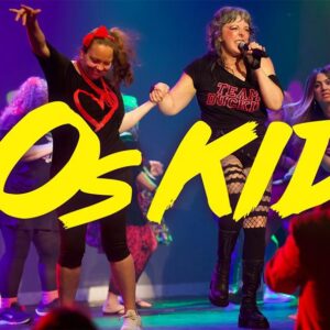 A lively performance with people in 80s-themed clothing, illuminated by colorful stage lights, with the text "80s KIDS" in bold yellow.