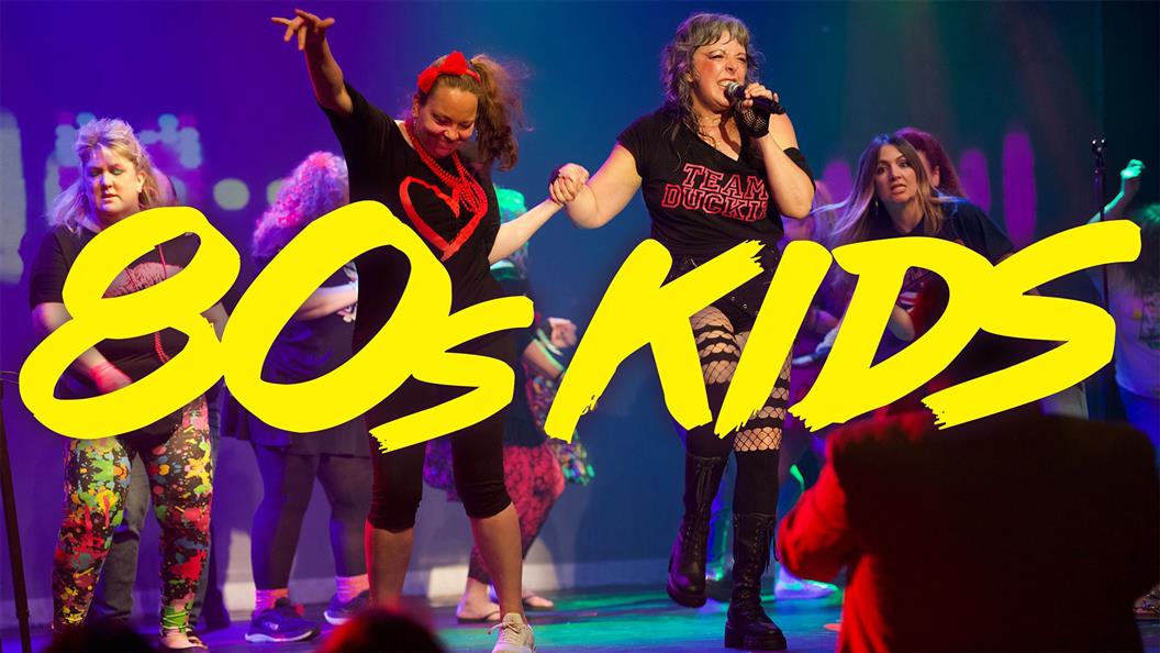 A lively performance with people in 80s-themed clothing, illuminated by colorful stage lights, with the text "80s KIDS" in bold yellow.