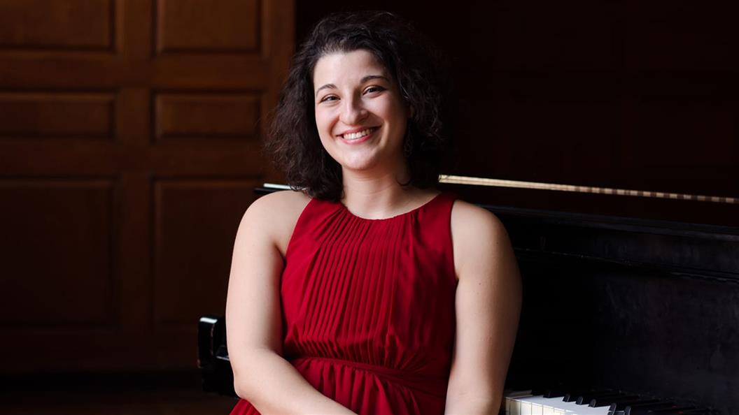 A person in a red dress sitting on a piano bench, smiling, with wooden paneling in the background.