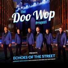 Six men in blue blazers stand on a city street at night with a large illuminated bridge in the background.