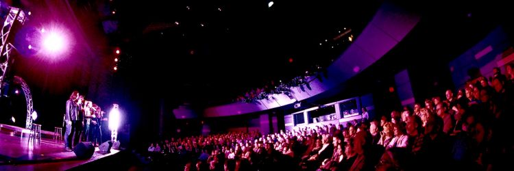 Panoramic view of a theater with performers on stage and an audience watching.