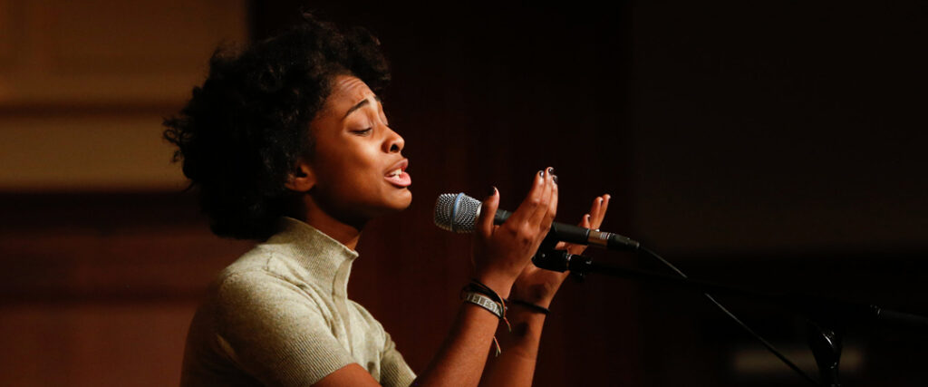 A person speaking into a microphone with an expressive gesture in a dimly lit setting.