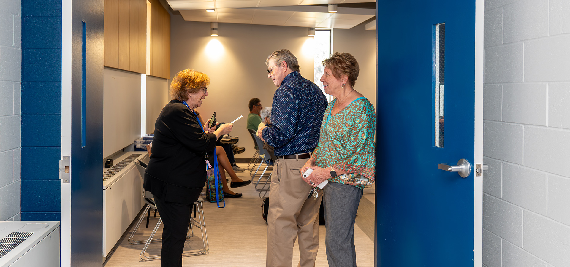 Three people converse in a hallway near an open blue door, with a waiting area and people seated in the background.