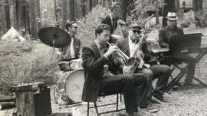 A jazz band performs outdoors in a forest, featuring a drummer, trumpet player, saxophonist, bassist, and keyboardist.