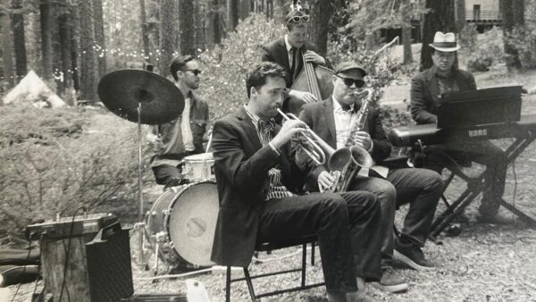 A jazz band performs outdoors in a forest, featuring a drummer, trumpet player, saxophonist, bassist, and keyboardist.