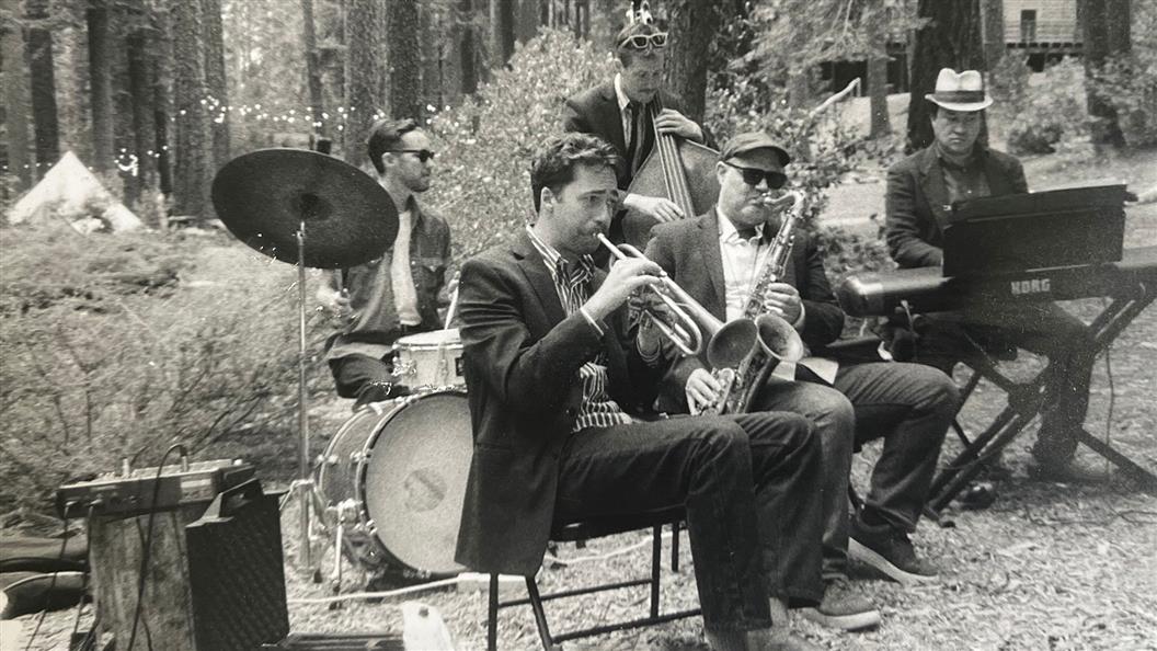 A jazz band performs outdoors in a forest, featuring a drummer, trumpet player, saxophonist, bassist, and keyboardist.