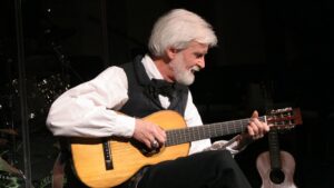 An older man with white hair playing a classical guitar on stage.