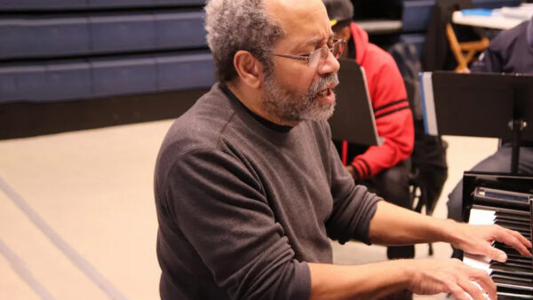 Person with gray hair and glasses playing a piano in a music practice room.