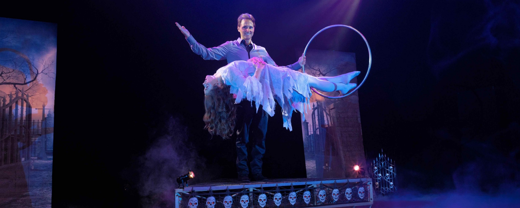 Theatrical scene with one performer holding a hoop behind another performer suspended horizontally in a tattered white costume above a skull-decorated platform.