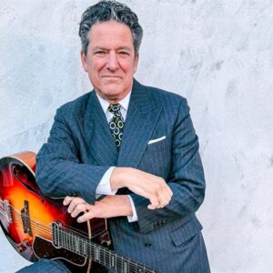 Man in a suit holding an acoustic guitar against a grey background.