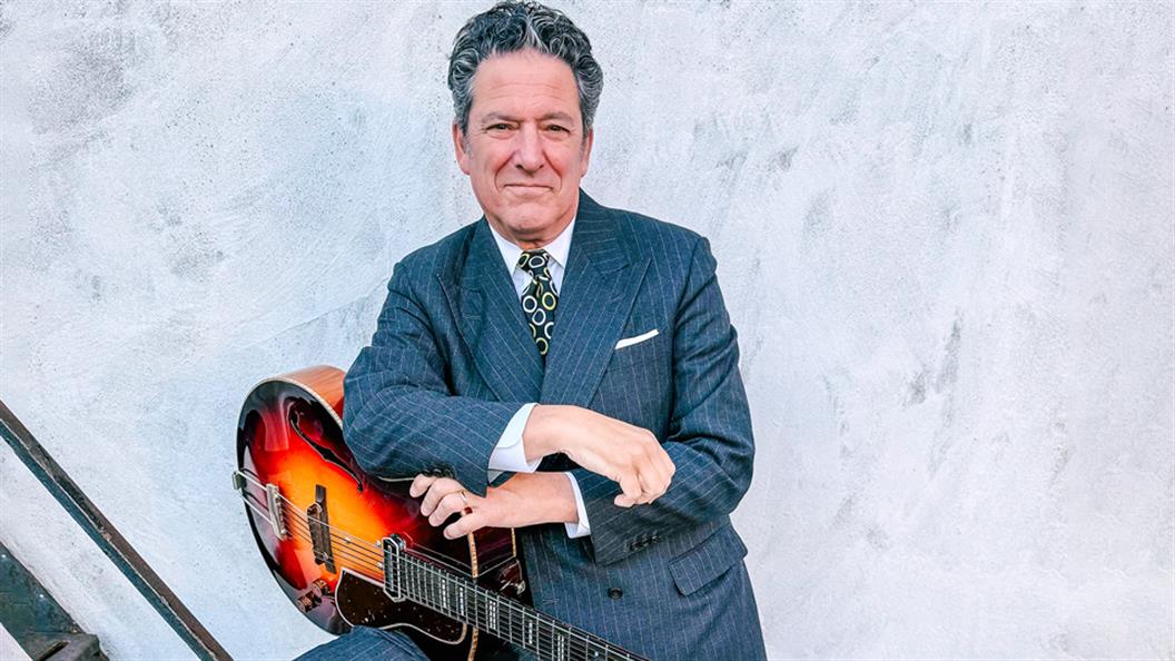 Man in a suit holding an acoustic guitar against a grey background.